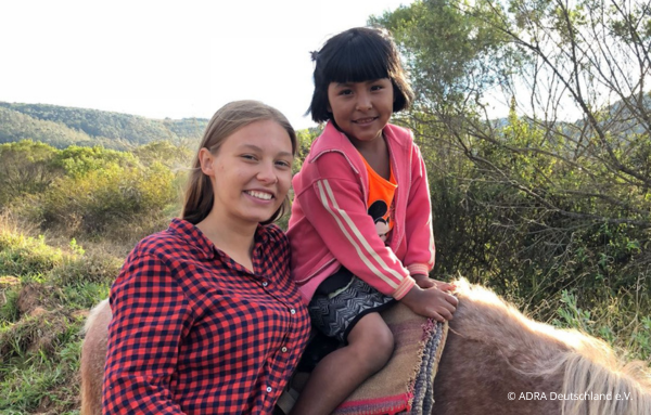 Die ADRAlive!-Freiwillige steht neben einem kleinen Mädchen, das auf einem Pferd sitzt, während beide in die Kamera lächeln.