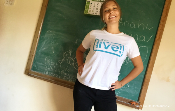 Eine ADRAlive!-Freiwillige steht vor einer kleinen Tafel und in einem Klassenraum im Kinderdorf „El Sauce“ in Bolivien. Sie trägt ein weißes T-Shirt mit dem Logo von ADRAlive! und grinst in die Kamera.