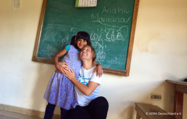ADRAlive!-Freiwillige steht vor einer kleinen Tafel in einem Klassenraum im Kinderdorf „El Sauce“ in Bolivien. Sie trägt ein weißes T-Shirt mit dem ADRAlive!-Logo. Neben ihr steht ein kleines Mädchen, welches sie umarmt und beide lächeln fröhlich in die Kamera