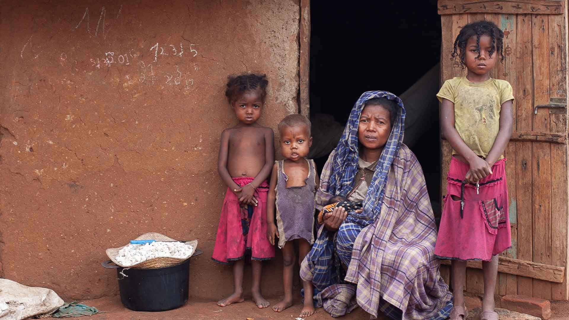 Eine Frau sitzt mir ihren unterernährten Kindern vor der Eingangstür, auf ihrem Schoß hält sie ein Säugling