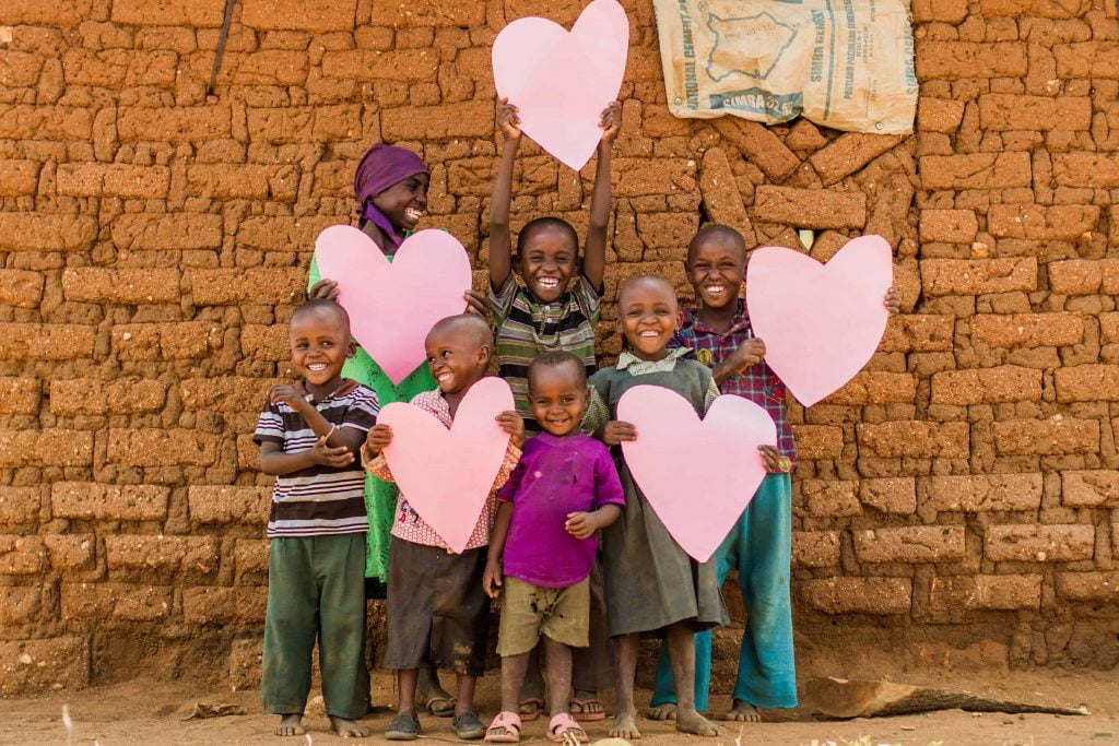 Eine Gruppe von lächelnden Kindern steht vor einer Mauer und halten rosa Herzen in den Händen