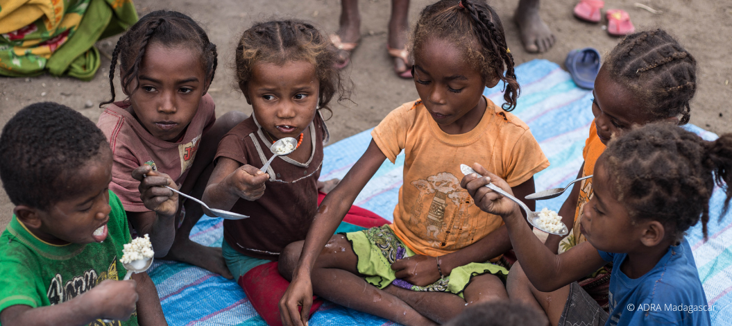 Eine Gruppe hungernde Kinder und Kleinkinder sitzt um eine Schüssel mit Brei gefüllt und teilt sich das Essen