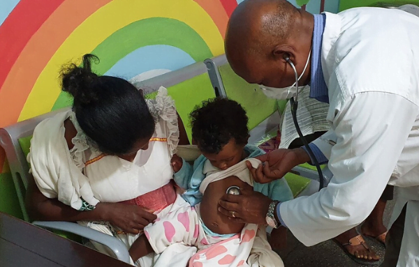 Ein Kleinkind sitzt auf dem Schoß der Mutter vor einer Regenbogenwand, während ein Arzt im weißen Kittel, das Kind mit einem Stethoskop untersucht