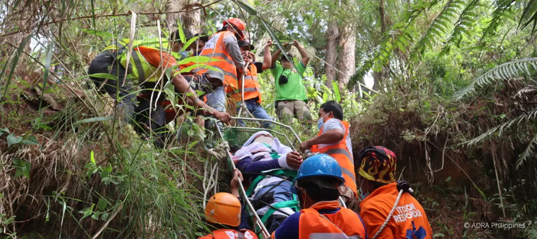 Ein Hilfstrupp im Wald transportiert einen Verletzen Menschen einen Hang zwischen den Bäumen hoch