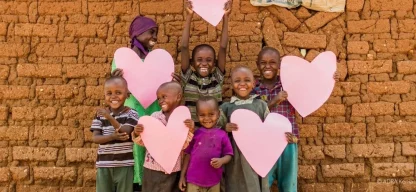 Lachende Kinder in Kenia halten große rosa Herzen vor einer Mauer aus Lehmziegeln