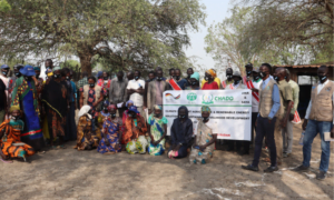 Eine Gruppe aus mehr als 20 Menschen, die im Südsudan Hilfsgelder von ADRA empfangen hat, sitzt vor Bäumen und stellt mit einem Schild das Projekt von ADRA vor
