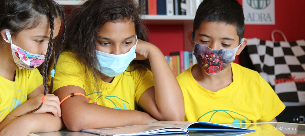 Drei Grundschulkinder in gelben ADRA-TShirts sitzen mit Mundschutz an einem Tisch und lernen