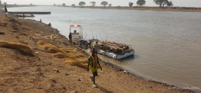 Ein Junge läuft am Ufer eines Flusses, dahinter ist ein Floss, mit dem Baumstämme und Holz transportiert werden