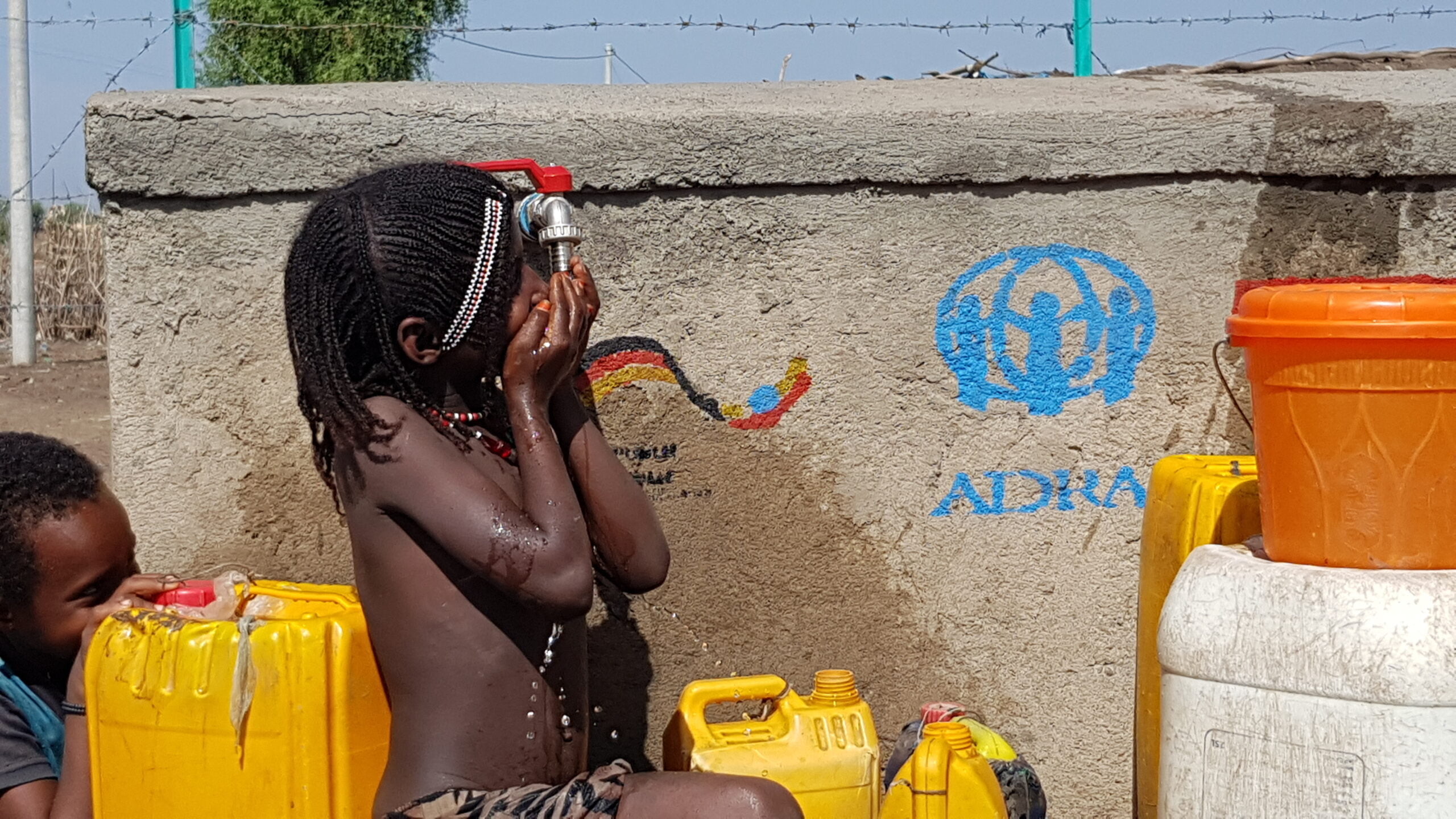 Kind trinkt an Wasserzapfstelle von ADRA, daneben stehen gelbe Wasserkanister