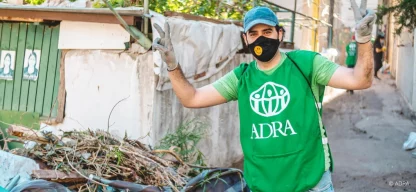 ADRA-Mitarbeiter in grüner Weste, blauer Basecap, Handschuhen und Smiley-Mundschutz hebt die Arme und zeigt das Peace-Zeichen