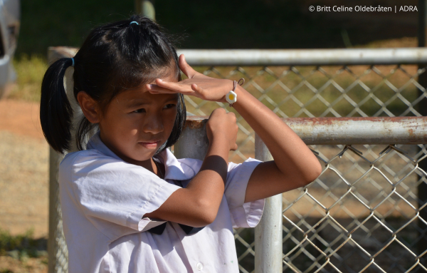 Ein Schulmädchen aus Thailand hält ihre Hand schützend an ihre Stirn, um sich vor dem blendenden Sonnenlicht zu schützen.