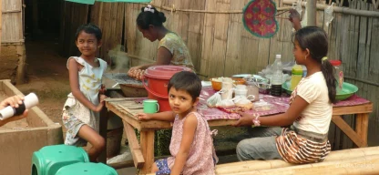 Kinder sitzen am Tisch und essen, eine Frau kocht