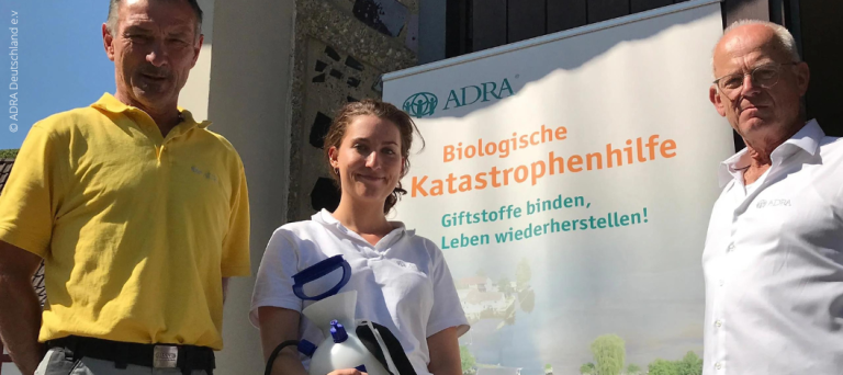 Hochwasserhilfe Effektive Mikroorganismen - Lächelnde Frau zwischen zwei Männern vor ADRA-Plakat hält Pumpe für Mikroorganismen