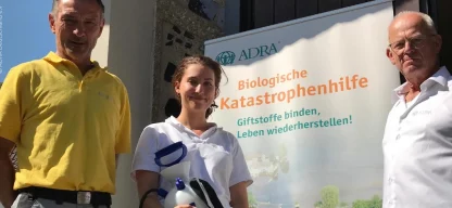 Hochwasserhilfe Effektive Mikroorganismen - Lächelnde Frau zwischen zwei Männern vor ADRA-Plakat hält Pumpe für Mikroorganismen