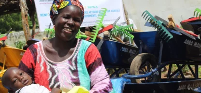 Frau mit Kind im Arm lächelt in die Kamera, hinter ihr Gartenwerkzeug und ein ADRA-Schild