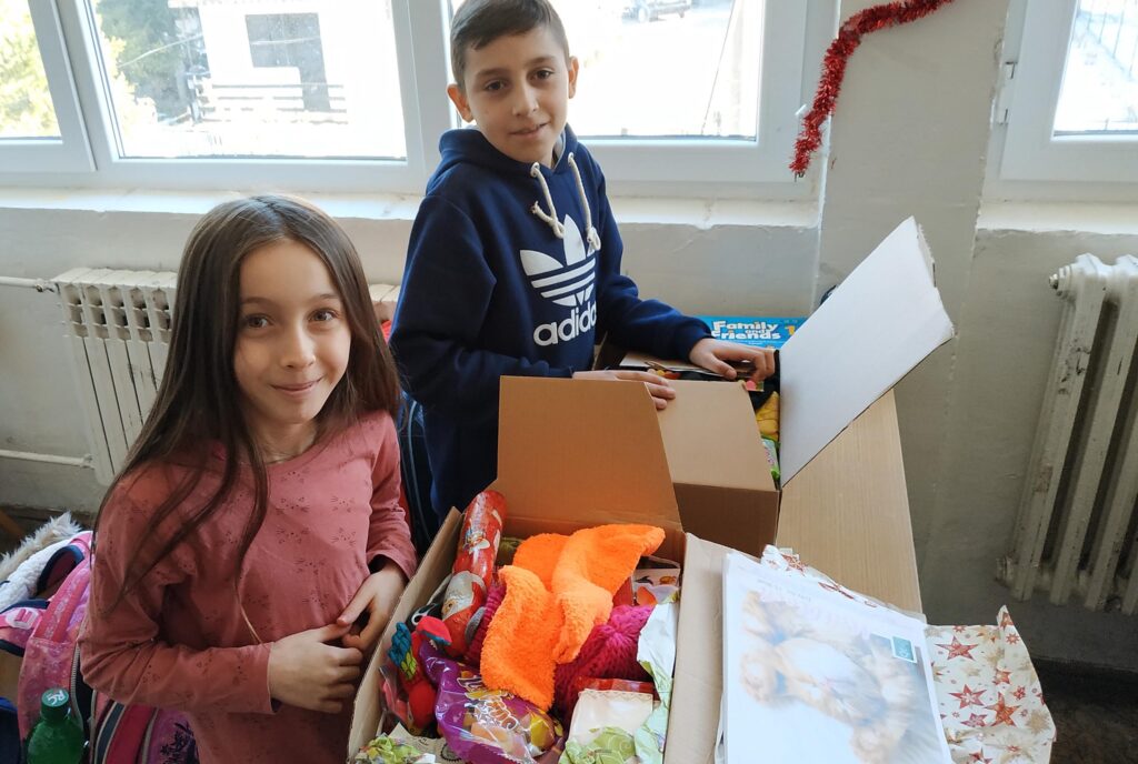 Ein junge und ein Mädchen packen gemeinsam KHK Pakete aus, welche aus Süßigkeiten, Spielzeuge, Kuscheltiere und Klamotten bestehen