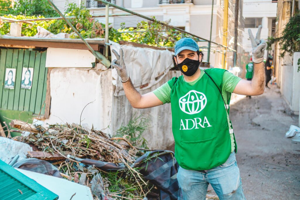 Ein Mitarbeiter von ADRA in einer grünen Weste, blauen Basecap, Handschuhen und einer Mund-Schutzbedenkung, auf der ein gelbes Smiley abgebildet ist streckt die Arme in die Luft und macht das Peace-Zeichen. Neben ihm ist ein Haufen mit Ästen