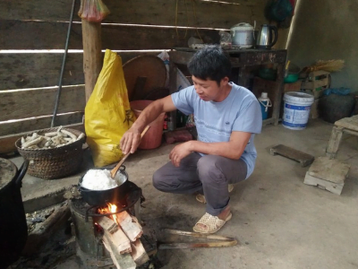 Ein Mann sitzt in einer Hütter in der Hocke, kocht Reis auf einer offenen Feuerflamme eines selbstgebauten Herds