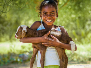 Ein kleines Mädchen hält eine Babyziege liebevoll in beiden Armen und lächelt in die Kamera.