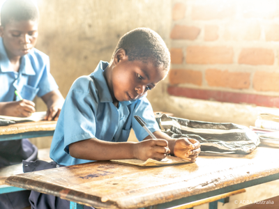 Ein Kind sitzt in einem Klassenraum an einem Schreibtisch und schreibt konzentriert in ein Heft. Das Bild verdeutlicht, wie Spenden für Bildung einem Kind die Chance geben, ein Schuljahr mit Schulgebühren, Uniform und Lehrmaterial abzuschließen.