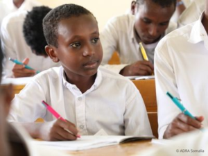 Kind sitzt in einem Klassenzimmer und schreibt konzentriert in ein Schulheft, weitere Kinder sitzen dahinter.