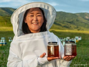 Eine Frau im weißen Imkeranzug steht auf einer Wiese und hält zwei Gläser Honig in der Hand, im Hintergrund stehen Bienenkästen.