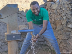 Ein Mann pumpt Wasser aus einer Handpumpe im Freien und wäscht sich lachend die Hände unter dem fließenden Wasser.