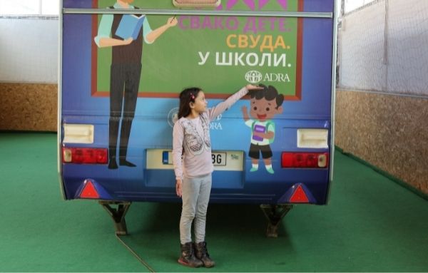Eine junge Schülerin steht vor einem mobilen Schulbus und zeigt mit der linken Hand auf das ADRA Logo