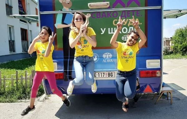 Drei fröhliche Schulmädchen springen vor der mobilen Schule in die Luft und zeigen mit beiden ihrer Händen das Peace-Zeichen