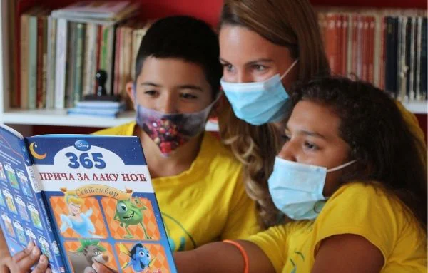 Zwei Schulkinder und eine Betreuerin mit Maske lesen ein Disney-Buch