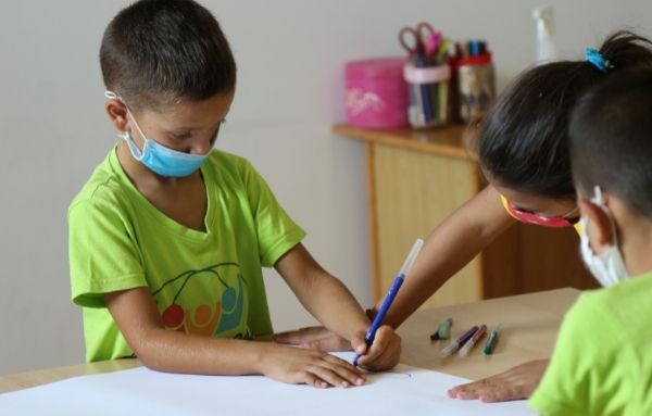 Drei junge Schüler sitzen an einem Tisch, tragen Mund-Schutz-Bedeckung und malen gemeinsam auf einem Papier