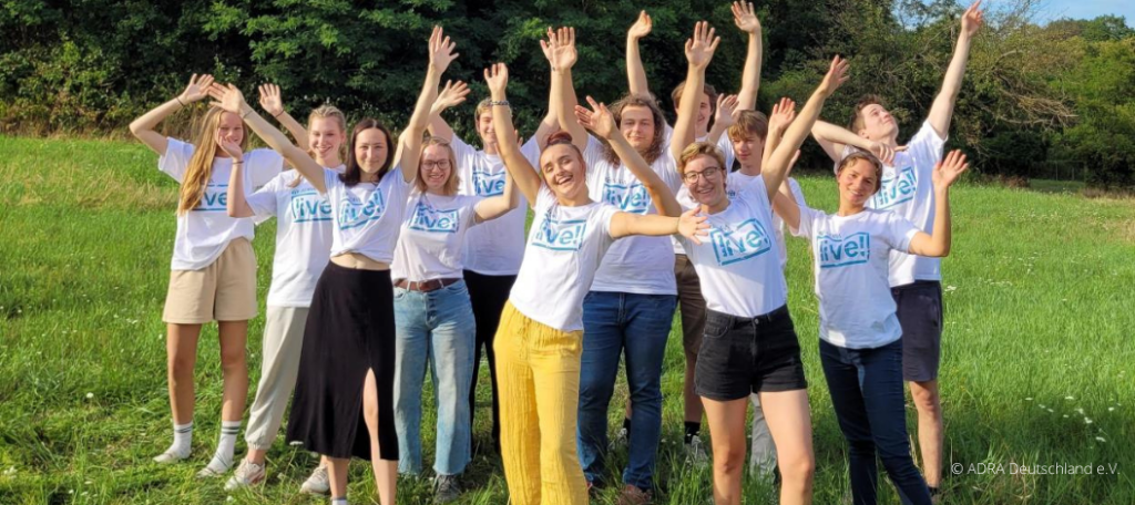 Eine lebendige Szene zeigt eine Gruppe von ADRAlive!-Freiwilligen in strahlend weißen T-Shirts, die sich auf einer üppig grünen Wiese versammelt haben. Mit einem begeisternden Lächeln auf ihren Gesichtern heben sie gemeinsam ihre Arme in die Luft, während das ADRAlive!-Logo stolz auf ihren Brustbereichen prangt