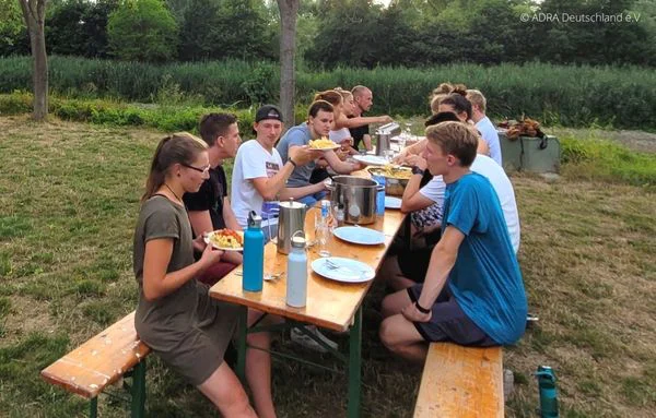 Junge Menschen sitzen an einer langen Tafel im Freien und essen zusammen. | © ADRA Deutschland e. V.