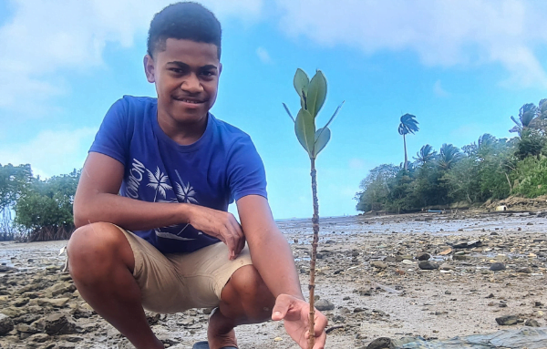 Ein lächelnder junger Erwachsener sitzt in der Hocke an der Küste von Fidschi und hält eine frisch eingepflanzte Mangrovenpflanze