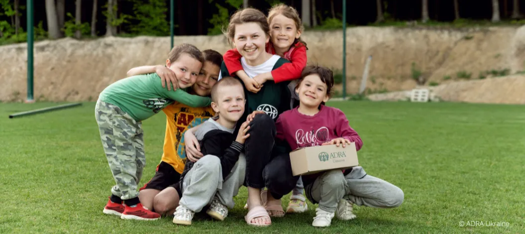 ADRA Mitarbeiterin sitzt mit glücklichen Kindern auf der Wiese, eines der Kinder hält ein Päckchen von ADRA auf dem Schoß.