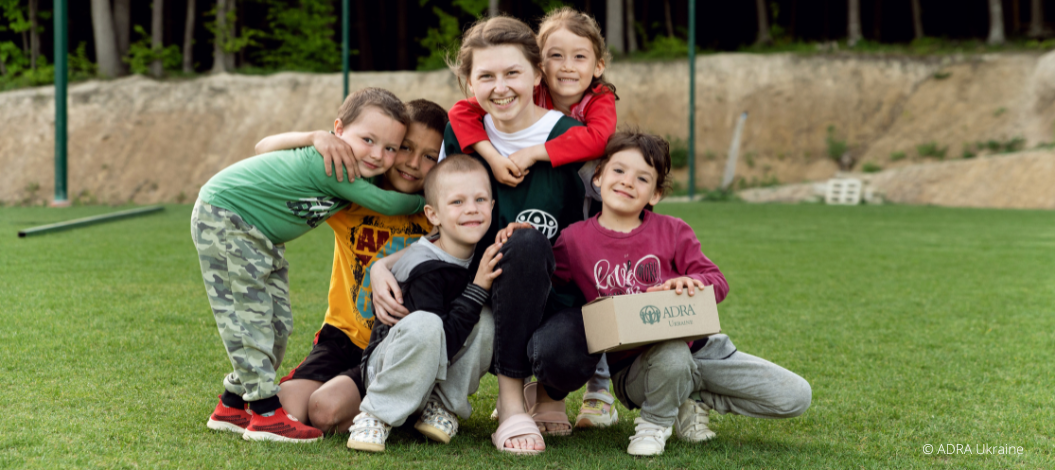 ADRA Mitarbeiterin sitzt mit glücklichen Kindern auf der Wiese, eines der Kinder hält ein Päckchen von ADRA auf dem Schoß.