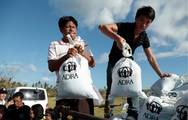 Zwei Männer aus Asien mit angestrengten Gesichtern, belagern die Lagerfläche eines Autos mit weißen ADRA-Säcken