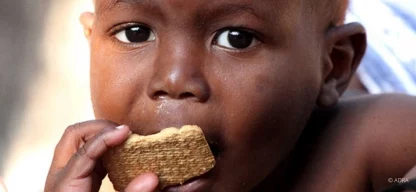 Ein kleines Kind hält einen Keks in der Hand und beißt hinein. Es blickt direkt in die Kamera. Der Hintergrund ist unscharf.