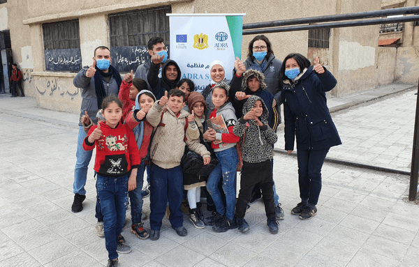 Eine Gruppe Schulkinder und vier Erwachsene mit Mundschutz-Bedeckung in Syrien, halten den Daumen nach oben und grinsen in die Kamera