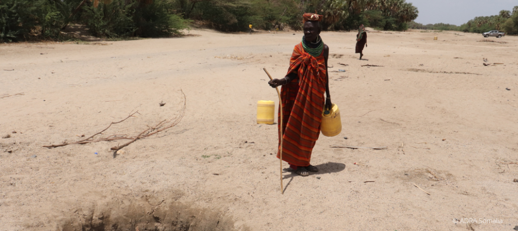 Eine Frau hält zwei gelbe Wasserkanister in den Händen. Vor ihr ist ein Brunnen im Boden, wo sie die Kanister auffüllt.