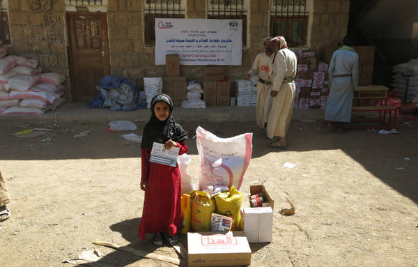 Ein junges Mädchen aus dem Jemen steht neben Nahrungspaketen und hält einen Zettel in der Hand. Im Hintergrund sind weitere Nahrungspakete an der Wand gestapelt und drei Männer.