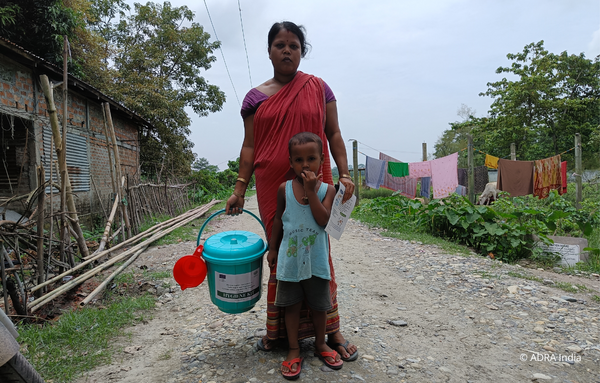 Eine Mutter aus Indien steht hinter ihrem Kind und trägt ein Hygiene-Set mit der einen Hand und eine Aufklärungsbroschüre über Hygiene in der anderen.