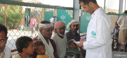 ADRA-Mitarbeiter im weißen Kittel spricht mit wartenden Männern und Kindern in einer Einrichtung im Jemen