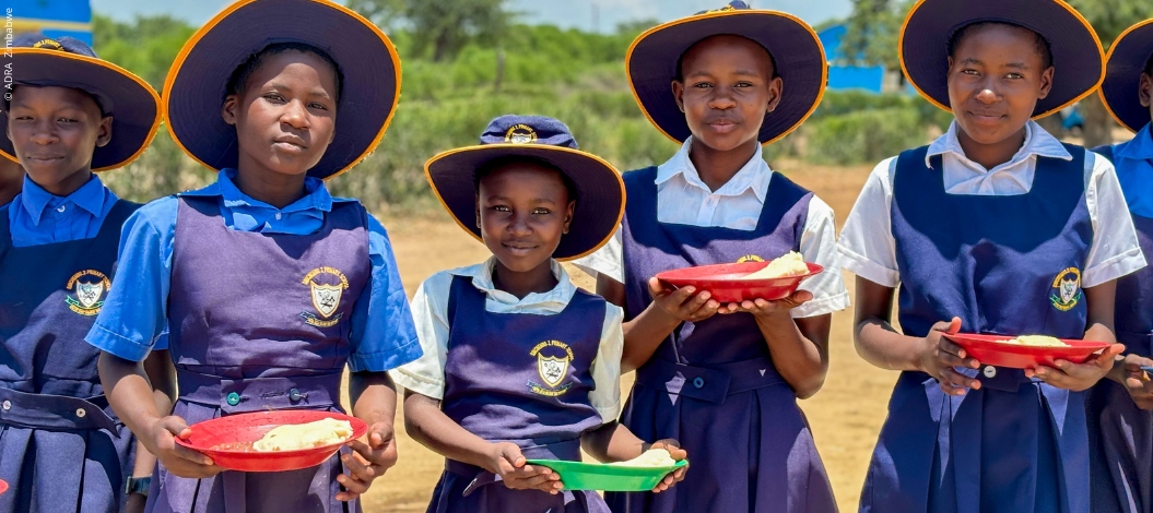 Eine junge Schulklasse aus Zimbabwe steht im Freien und hält Teller mit Essen in den Händen, während sie in die Kamera lächeln.