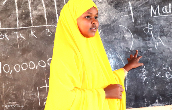 Khadra Muse, eine Schülerin aus Somalia welche schreiben und lesen gelernt hat, steht vor einer Schultafel auf welche sie ebenfalls mit ihrer Hand zeigt.