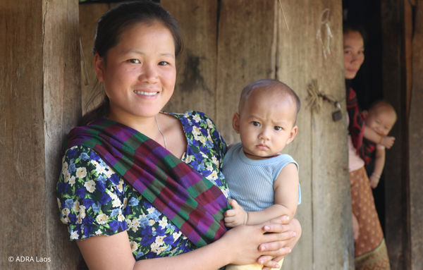Eine Bäuerin aus Laos, hält ihren Säugling im Arm und lächelt freundlich in die Kamera. Im Hintergrund ist eine weitere Frau mit einem Säugling im Arm.