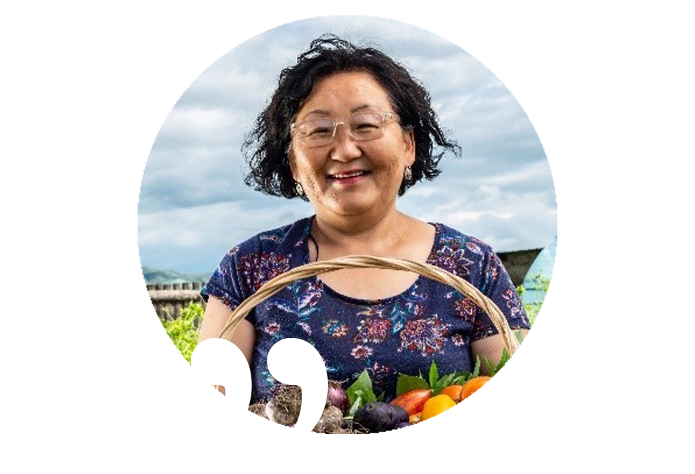 Eine Frau steht mit einem gut gefüllten Gemüsekorb lächelnd vor einem Gewächshaus auf einem Feld