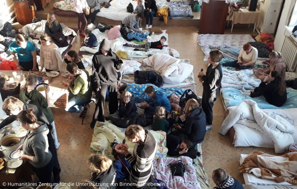 eine Gruppe von Menschen, die um einen mit Betten gefüllten Raum des humanitären Zentrums für die Unterstützung von Binnenvereinen herumstehen und sitzen.
