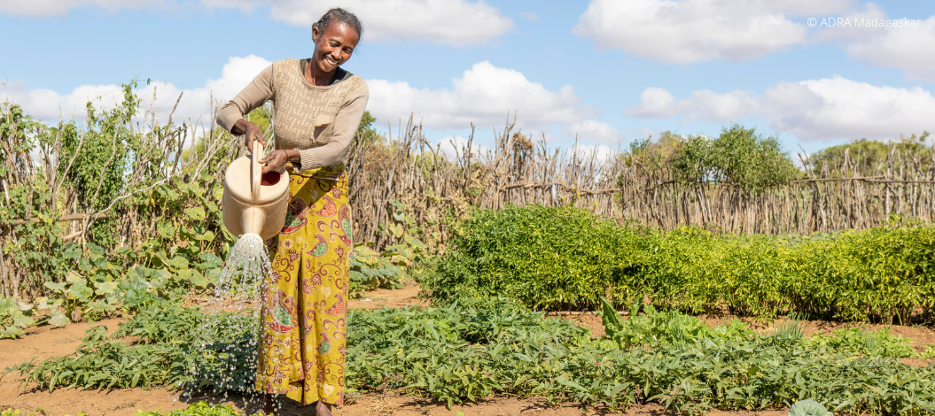 Eine lachende Frau hält mit beiden Händen eine Gießkanne und bewässert ihre Bio Ernte.