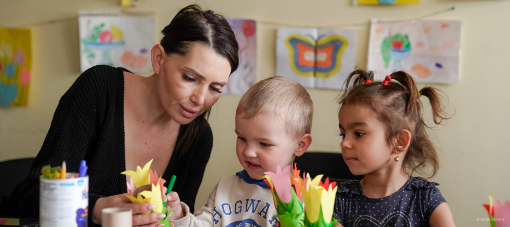 Eine Frau zur Linken sitzt mit zwei Kindern am Tisch und malt mit ihnen.